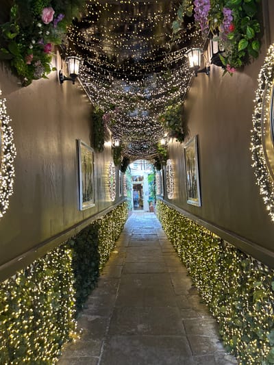 An outdoor alleyway is adorned with festive lights. The left side is a wall decorated with greenery and cascading fairy lights, punctuated by lamp fixtures. The right side features a stone wall with greenery and string lights along the base. Mirrors are installed along the ceiling and a stone pathway leads to an illuminated doorway at the end of the alley. Fairy lights encircle the ceiling at both ends of the alley.