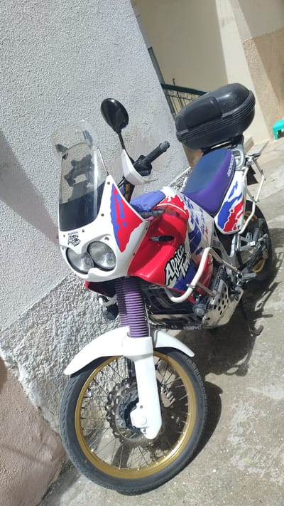 The image shows a white Honda Africa Twin motorcycle with red, blue, and purple decals parked on a concrete surface next to a textured, light-colored wall. The motorcycle has a black top case, a clear windscreen, and gold-colored rims on its wheels. The front fork gaiters are purple. The background suggests an outdoor urban environment, possibly in Νέα Μηχανιώνα, Greece, with a building and some shadows visible. There are no people in the image.