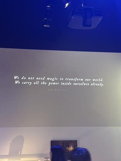 The image depicts a projector screen displaying a quote attributed to J.K. Rowling: "We do not need magic to transform our world. We carry all the power inside ourselves already." The quote is in white, serif font and centered on the screen. Above the screen, a projector is mounted on a dark blue ceiling. The lower portion of the image shows a blurred view of the audience, with the top of heads and shoulders visible, suggesting a presentation or screening event. A shadow of a figure is cast on the wall to the left of the frame. The scene appears to be indoors, possibly in a lecture hall or cinema, in the city of Three Rivers, United Kingdom.