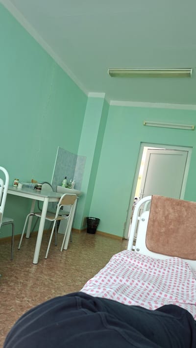 This is a photograph of a hospital room, likely in Nytva, Russia, as indicated by the location context. The room is painted a light green, with white trim. Fluorescent lights are mounted on the ceiling. The room appears to be a ward, with a hospital bed in the foreground covered with a pink and white checkered blanket and a brown blanket. The bed is made of white metal. To the left of the bed, there is a sink area with a tiled backsplash and bottles of liquid, suggesting hygiene products. A white table with chairs is positioned near the sink. A black trash can is visible in the corner. The floor is covered in a patterned linoleum. A doorway with a white door is visible in the background, leading to a brightly lit area, possibly an exterior or hallway. A crack is visible in the wall near the floor. The image is taken from a person's perspective, with their dark clothing visible in the lower right corner, suggesting they are either a patient or a visitor. The lighting indicates it is daytime. No text is visible in the image.
