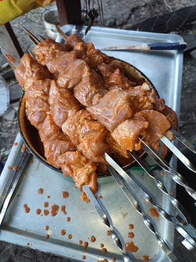 In Voskobiinyky, Ukraine, raw, marinated meat pieces are skewered onto metal and wooden sticks, arranged in a brown circular bowl. The meat appears to be coated in a reddish-brown marinade with visible spices. The bowl sits on a rectangular metal tray, which has several brown liquid droplets scattered across its surface. The background shows an outdoor setting with the ground and some wooden elements. The scene suggests preparation for cooking, likely barbecue or shashlik.