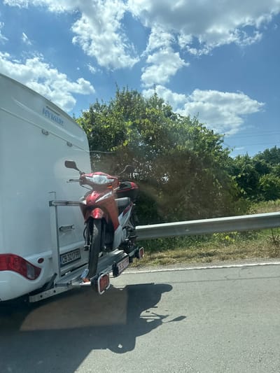 This media file captures a scene of a white camper van with a red and silver scooter mounted on a rear rack. The van has "HYMER" written on its side. The scooter's license plate is visible with the text "CB 3217 PP".
The scene appears to be on the side of a road, with a grassy verge and trees lining the background. A metal pole, possibly a roadside barrier or utility pole, runs vertically along the right side of the image. The road surface is asphalt.
The time of day is suggested to be daytime by the bright sunlight casting shadows of the scooter and its rider onto the road. The sky is blue with scattered white clouds, indicating fair weather.
The image is taken from a slightly elevated perspective, looking down and to the side, as if from another vehicle traveling alongside or behind the camper van. The overall impression is one of travel and outdoor adventure. The location context provided is Asparuhovo, Bulgaria.