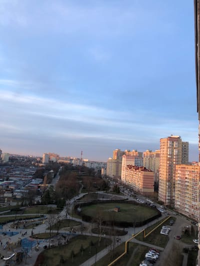 The image showcases a cityscape under a predominantly clear, pale blue sky. Towering residential buildings dominate the upper portion of the frame, transitioning into smaller structures lower down. A green park with pathways is visible, adjacent to a road with parked cars. The overall scene conveys an urban environment on a clear day.