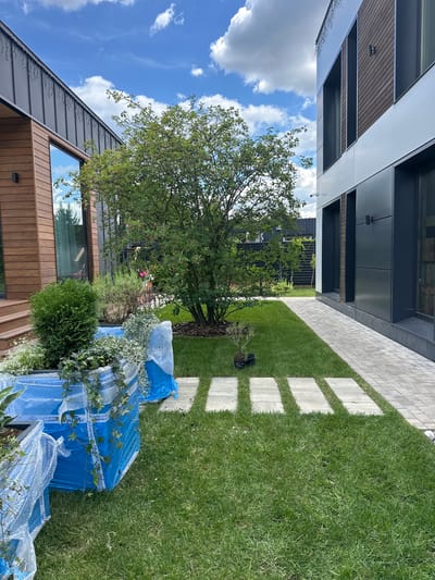 The image captures a well-maintained garden area outside a modern house in Лапино Град Кп, Russia. The scene is set during the daytime, with a clear blue sky dotted with white clouds, suggesting pleasant weather.
The primary subjects include elements of the house and its surrounding landscape. The house features contemporary architecture with a combination of dark grey metallic siding, wooden accents, and large windows. A paved walkway made of light grey rectangular stones runs through a lush green lawn, interspersed with square concrete stepping stones. A large, mature tree with vibrant green foliage stands as a central feature in the garden, casting some shade. In the foreground, several items are wrapped in blue plastic sheeting and bubble wrap, suggesting they are new materials or stored items for the property. Some greenery, including a small evergreen bush and ivy, are also visible.
There are no people visible in the image, and the activity appears to be static, likely related to ongoing landscaping or home improvement. The overall mood is serene and peaceful, reflecting a well-kept outdoor environment. The visible text on the blue wrapping appears to be in Cyrillic and includes phone numbers such as "+7 (926) 702 51 16" and a website "www.walmi.ru", which might indicate the supplier or manufacturer of the wrapped items. The overall impression is one of a modern, well-appointed property in a suburban or semi-rural setting.