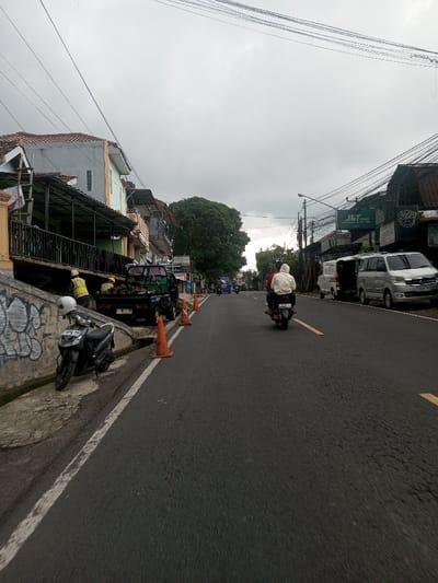 The image depicts an urban street scene in Kandapola, Sri Lanka, under an overcast sky. The asphalt roadway features a solid white line marking the left shoulder and dashed yellow lines in the center.
On the left side of the street, in the foreground, a concrete retaining wall exhibits black graffiti. A black scooter with a white helmet resting on its seat is parked on the sidewalk next to this wall. Behind the scooter, a green pickup truck is parked by the curb, its bed loaded with dark material. Two individuals are partially visible near the truck; one wears a yellow vest, and another wears a light-colored shirt, both appearing engaged in an activity by the truck or the elevated structure above it. Three orange traffic cones are positioned on the street adjacent to the curb, extending past the pickup truck. Beyond this, multi-story buildings with tiled roofs and balconies line the street, some featuring awnings. A large tree with dense green foliage is prominent mid-ground on this side.
On the right side of the street, two individuals are riding a motorcycle away from the viewer. The passenger wears a white hooded garment, and the driver wears a red shirt. Further down, two silver-colored vans are parked on the roadside. Buildings with commercial signage, including "J&T Cargo" and "SENA," are visible on this side. A street light pole stands near the parked vans.
Overhead, numerous utility lines crisscross the scene against the grey, overcast sky. The overall lighting is diffuse, consistent with an overcast day. Other vehicles and indistinct figures are visible in the distance, indicating general street activity.