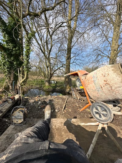 The image depicts a first-person perspective of an outdoor work site adjacent to a body of water in Codford, United Kingdom, under a clear sky. In the immediate foreground, the lower legs of an individual are visible, dressed in dark, dirt-covered trousers and light-colored work boots extensively splattered with mud. The individual appears to be seated or resting on a flat, soiled surface. To the left of the legs, a yellow and black cable reel or portable generator is positioned on the ground.

The midground features a narrow body of water, consistent with a stream or pond, flanked by a muddy bank strewn with broken bricks, small pieces of wood, and other construction debris. On the left bank, a long-handled tool, possibly a shovel, leans against a tree trunk, with an orange chainsaw resting on a wooden plank nearby. On the right, a weathered orange cement mixer, featuring a large drum and black wheels, stands prominently on uneven ground. Several wooden pallets are visible behind the mixer, near the water's edge.

The background reveals a line of leafless deciduous trees against a bright blue sky with sparse white clouds, indicative of late winter or early spring. Beyond the trees and water, an uncultivated field or wooded area extends. No other individuals or specific interactions are discernible. The scene implies a temporary pause in active construction or land-clearing operations.