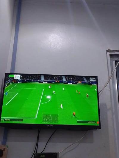 A television displays a soccer video game with two teams, ESP (Spain) and MAR (Morocco). The score is ESP 0, MAR 2. The time is 63:30. The game is being played on a green field with white lines. There are several players on the field, some in white uniforms and some in orange and red uniforms. The game is being played in a stadium with a crowd visible in the background. The TV is mounted on a light gray wall. To the right of the TV is a white electrical cable. Below the TV is a black set-top box with a digital display showing "01 01". The overall scene suggests a living room or entertainment area. The location is identified as Abeere, Nigeria.