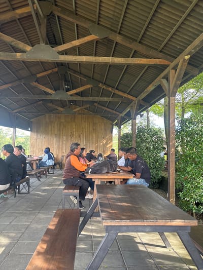The scene depicts an outdoor, covered dining or social area situated in Tamiajeng, Indonesia. The overhead structure consists of a dark corrugated roof supported by a complex network of dark wooden beams, rafters, and black metal plates securing the structural connections. Several industrial-style dome light fixtures are suspended from the ceiling beams. The ground is paved with light gray square tiles. A vertical wall constructed from light-colored wooden planks forms the rear boundary on the left, while the right side of the structure remains open, revealing dense green foliage, bushes, and trees. A distant, lighter green landscape is visible beyond the wooden wall to the far left.
Approximately 15 to 20 adults are present, seated at multiple rectangular dark wooden tables, each paired with matching dark wooden benches, all featuring black metal support frames. In the middle-left foreground, a man with graying hair, wearing an orange vest over a brown shirt and dark pants, is seated opposite a woman in a pink top, both engaged in conversation. A black backpack rests on their table. Further left, several individuals are seated, including one prominent figure in a black t-shirt and glasses. In the middle-right, four men are seated at a table; one in a dark t-shirt and light blue jeans holds a mobile phone, while another in a black t-shirt observes the table. Mobile phones are visible on multiple tables. The individuals are primarily seated, conversing, and interacting with their mobile devices. An empty dark wooden table with an accompanying bench set occupies the immediate foreground.
