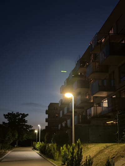 The image depicts a nighttime scene on a paved walkway leading towards a dark, leafy tree on the left. The walkway is illuminated by a streetlamp on the right, which is a modern design with a tall, slender pole. Adjacent to the walkway, on the right, is a well-maintained lawn bordered by neatly trimmed green bushes. In the background, a multi-story modern building with numerous balconies is visible, constructed with concrete and glass. The building's balconies have glass railings, and some windows are lit from within. The sky is a deep, dark blue, indicating late evening or early night, with faint clouds present. There is a green, pixelated light or sign in the sky. This scene is characteristic of the Bystrzyca district in Lublin, Poland, specifically an area with modern residential buildings and green spaces. No people are present in the image.