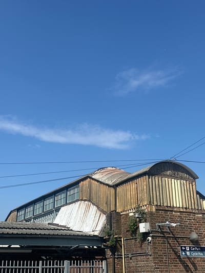 The image, when viewed upright, displays an architectural detail of a building under a bright blue sky. The left side of the frame is dominated by a clear, vibrant blue sky, featuring a few wispy, elongated white clouds. Several thin, dark wires stretch horizontally across the upper portion of the sky, connected to a structure on the right.
The right side of the photograph features a weathered industrial-style building. Its roof is characterized by a series of curved, corrugated metal sheets, showing signs of age with various tones of grey, brown, and hints of rust. Below this, another section of the building reveals a façade with multiple small-paned windows, suggesting a clerestory or sawtooth roof design that allows light into the interior.
A reddish-brown brick wall forms a prominent part of the building's exterior. Mounted on this wall are several fixtures: two security cameras, a grey utility conduit, a white electrical box, and a grey speaker or light. Small patches of green vegetation are growing directly from the brickwork in various spots.
A notable detail on the brick wall is a blue directional sign with white text and symbols. It reads "P Car Park" with an arrow pointing left, and "Cycle Hub" with an arrow pointing diagonally down-left. Below this, a dark, grid-like metal structure is visible in the lower right, possibly part of a walkway, bridge, or station platform.
The scene is well-lit by direct sunlight, casting clear shadows and indicating it is daytime. There are no people visible, and no specific activity is taking place. The presence of "Car Park" and "Cycle Hub" signs, along with the industrial architecture and the given location context of "Waverley, United Kingdom," strongly suggests this is part of or adjacent to Edinburgh Waverley railway station.