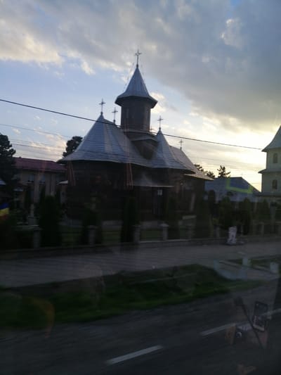 A wooden church with multiple steeples and crosses stands against a cloudy sky. In the foreground, a street and grassy area are visible, reflecting the scene. To the left, a building with a red roof and visible windows is situated behind a fence and some trees. The overall impression is of a serene, possibly rural, religious establishment.