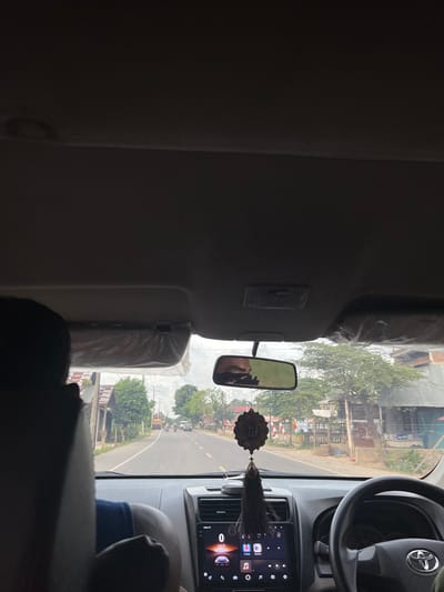 The image depicts the interior of a vehicle traveling on a road in Matangkumbang, Indonesia. The perspective is from the back seat, looking forward. In the foreground, the back of a person's head is visible on the left. The dashboard of the car features a touchscreen infotainment system displaying icons for various applications and a speedometer indicating 0 km/h. A steering wheel is visible on the right, with a Toyota logo in the center. A rearview mirror hangs from the ceiling, reflecting the face of a person in the driver's seat. A decorative Islamic ornament with tassels dangles from the rearview mirror. The road ahead is lined with houses, shops, and trees. Several motorcycles and cars are visible on the road, suggesting moderate traffic. The sky is overcast, and flags are displayed along the roadside. The overall scene captures a typical journey through a semi-urban or rural area in Indonesia.