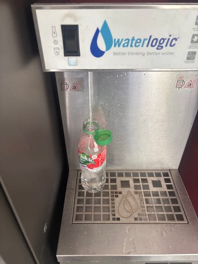 This image captures a close-up of a Waterlogic water dispenser in an indoor setting in Lièpvre, France. A clear plastic 50 cl "Volvic Juicy" bottle with a red and white strawberry-themed label is positioned on the stainless steel drip tray, actively being filled with water from the central dispensing nozzle. The bottle's green flip-top cap is open and resting on the bottleneck. The dispenser's top panel is white, featuring the "waterlogic" logo with blue teardrop elements and the slogan "Better thinking. Better water." A black toggle switch and a small blue button are visible on the left side of this panel, alongside icons for a bottle, power, a glass, and electricity. The stainless steel face of the dispenser shows water droplets and features two distinct warning labels, each depicting a steaming cup and a hazard triangle, positioned to the left and right of the dispensing area. The drip tray is a metal grate with a subtle, perforated double-teardrop design in its center. The background on the left is a dark, solid surface, while a dark red surface is visible on the right. No individuals are present in the frame.