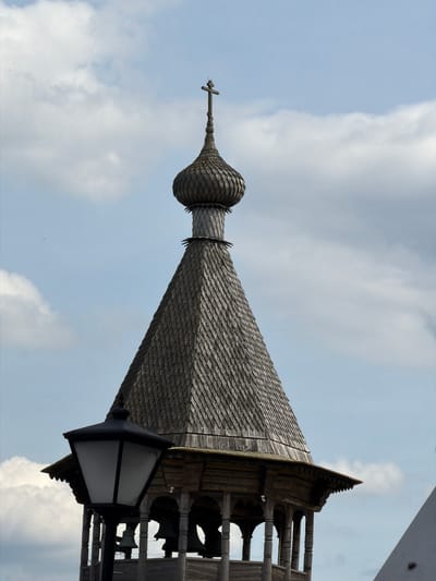 In Nevskii Parkleskhoz, Russia, a close-up shot captures the upper section of a wooden bell tower topped with a distinctive onion dome and a cross. The tower is covered in weathered wooden shingles, giving it a textured, dark gray appearance. Below the conical roof of the tower, an open wooden structure is visible, featuring a series of arched supports. Several dark, bell-shaped objects, presumably bells, are hanging within this lower section. In the foreground, a black lamppost with a lantern-shaped head stands to the left, its white light panel partially obscuring the bell tower. The background consists of a pale blue sky with scattered white clouds.