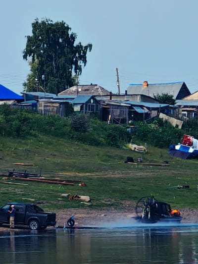 The image depicts a waterfront scene in the village of Pogodaevo, Russia, on what appears to be a clear day. In the foreground, the calm, reflective surface of a body of water dominates the lower portion of the frame.
On the shore to the left, a dark-colored pickup truck is parked, with a person wearing a white cap and light-colored pants standing near the open driver's side door. Adjacent to the truck, a tire lies on the ground next to a water-level launch area. Further out on the water, a black hovercraft is in motion, emitting a plume of mist and spray as it travels.
The shore itself is a grassy slope, with various objects scattered on it. To the left, a pile of logs and wooden planks are visible. Further up the slope, a collection of various structures are situated, appearing to be a mix of small houses, sheds, and outbuildings. These buildings are varied in construction, with some featuring tin or corrugated metal roofs, and others appearing to be more rustic wooden structures. A tall, leafy tree stands prominently behind these buildings.
In the mid-ground and background, the landscape continues with more greenery and the structures of the village. Power lines and a wooden utility pole are visible in the upper portion of the image. The sky is a pale blue, with no clouds apparent. The overall impression is of a functional, somewhat rural, waterside settlement.