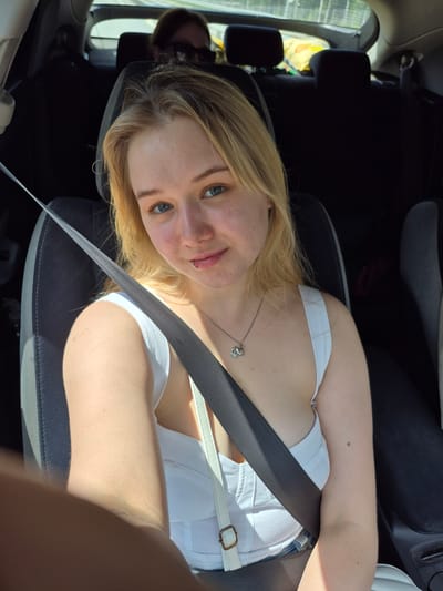 A young woman with blonde hair and blue eyes is seated in the front passenger seat of a car, wearing a white top. She has a lip piercing. A dark gray seatbelt is fastened across her chest. The background shows the interior of the car, with the back seats visible and two other people, one in the back wearing sunglasses and the other partially obscured. Yellow bags are in the back seat. The scene appears to be during the daytime. The specific location in the city of Сарс, Russia cannot be determined from the image.