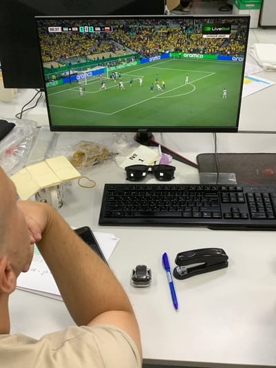 The image shows a person sitting at a desk with a computer monitor displaying a soccer match. The score on the screen reads "UZB 1 2 COL" with 88:37 elapsed time. The live ball graphic indicates it is a direct broadcast. The desk is cluttered with office supplies, including a keyboard, mouse, stapler, pens, sunglasses, and a stack of paper. A person's arm and shoulder are visible in the foreground, suggesting they are viewing the monitor. The background shows office equipment and possibly a window. The mention of Urtaaul, Uzbekistan, in the prompt is not supported by any visual evidence in the provided image.