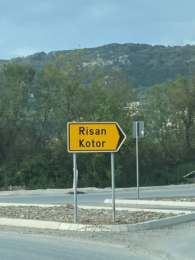 The image shows a yellow and black road sign indicating the directions to Risan and Kotor in Kavač, Montenegro. The sign is mounted on metal poles beside a road with a concrete barrier. Behind the sign is a hillside covered in green vegetation, and a blue sky is visible above. A smaller rectangular sign is situated above the direction sign. The photograph is taken from a roadside location.