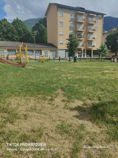 The image depicts an outdoor scene in Sondrio, Italy, on June 5, 2026, at 16:32. The foreground consists of a large, uneven grassy area with visible patches of dry, light-brown grass and areas of greener growth.
In the midground, a children's playground structure with a red slide, yellow framing, and blue safety netting/bars is visible on the left. A child, dressed in a dark shirt and shorts, is standing near the base of the playground structure. To the right of the playground, a one-story building with a tiled roof, multiple windows, and a beige facade is partially visible. Further to the right and slightly behind, a prominent multi-story residential building, painted light beige, features numerous windows with white external blinds (some closed) and multiple balconies with dark railings. Laundry is visible drying on one of the balconies on the third floor from the bottom.
In front of the residential building, a paved area or sidewalk is visible, along with a grey cylindrical trash receptacle and a light-colored bicycle parked near a closed corrugated metal door. Several trees are planted in front of the buildings, and a street light pole stands between the smaller building and the apartment block. On the far right, a woman with light-colored hair and clothing is seated on a bench, with a beverage container next to her.
The background features densely forested mountains under a sky with scattered white clouds. The entire scene is illuminated by daylight, casting shadows consistent with the specified time of day. Overlays on the image include "TIKTOK PRO:KRi120918" in the bottom left corner and "The decisive Moment" in script in the bottom right corner.