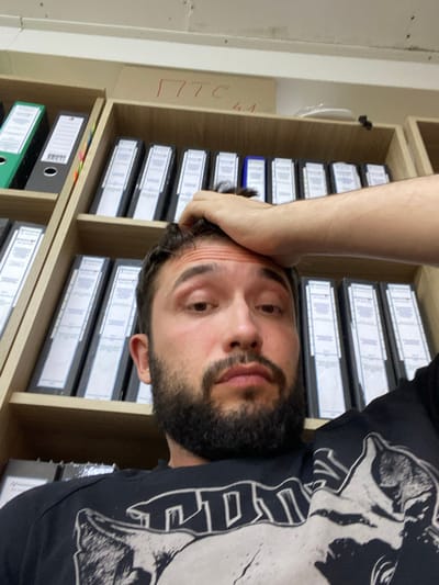 A man with dark hair and a beard is leaning back with his hand on his forehead, looking towards the camera. He is wearing a black t-shirt with a graphic design. Behind him, shelves are filled with binders and folders. The room appears to be an office or storage area. The text "ПТС 41" is written in red on the top shelf. The location is specified as Luzhitsy, Russia.