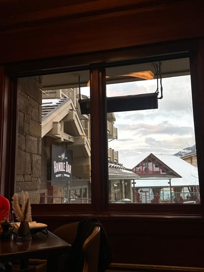 The image presents a view from inside a building, looking outward through a dark wooden window frame. The location is Teton Village, United States of America.
In the immediate foreground, to the lower left, a dark polished wooden table is partially visible. It holds a small, clear glass vase containing dried, light brown pampas grass-like decorative reeds, and a dark, cylindrical condiment dispenser (likely a pepper mill). A light-colored, possibly leather, upholstered chair with a dark garment draped over its back occupies the lower-right foreground. The interior features dark wooden paneling below the window.
The exterior scene reveals a snowy resort village under an overcast sky with some lighter patches. Directly outside, multiple buildings constructed with stone and wood feature snow-dusted gabled roofs and exposed balconies. A prominent black sign, suspended from an overhead structure, reads "THE HANDLE BAR WHISKEY & PATIOS" in white lettering. Several black, rectangular outdoor heaters are visibly suspended from architectural overhangs. Further into the midground, another building with a snow-covered gabled roof and a large window with distinct red framing is discernible. Bare tree branches are visible on the far right. No identifiable individuals or specific actions are observed.