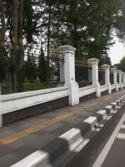 The image captures a roadside scene, depicting a long, decorative fence running parallel to a gray paved sidewalk. The fence features regularly spaced white pillars with intricate caps, connected by black wrought iron panels topped with gold-colored, spiky finials. The fence's lower section consists of a white concrete wall above a dark, rough-hewn stone base. The sidewalk includes a prominent yellow tactile paving strip. Adjacent to the sidewalk is a curb, painted with alternating black and white stripes, bordering a blurred road surface. Behind the fence, a verdant green lawn extends into the midground, populated by numerous tall, slender trees, some resembling conifers, under an overcast sky. The entire image exhibits significant motion blur, consistent with being captured from a moving perspective. No individuals are visible. The location is specified as Chagzam_Pam, Bhutan.