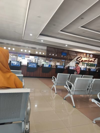 This media file captures the interior of a government office, likely a Samsat (Vehicle Registration and Identification) office in Barengkrajan, Indonesia. The scene shows a waiting area with rows of silver metal chairs, some occupied and some empty. In the foreground, a person wearing an orange hijab is seated, facing away from the camera.

Behind the waiting area, a counter is visible with several service windows. Signs above these windows indicate various services offered, such as "PENDAFTARAN 1 (5 TAHUNAN & BBN 2)" (Registration 1 - 5 Years & Vehicle Tax 2), "LAYANAN DUPLIKAT STNK" (Service for Duplicate Vehicle Registration Certificates), "MUTASI KELUAR" (Outgoing Mutation), and "PENETAPAN PAJAK" (Tax Determination). A large sign in the background reads "KANTOR BERSAMA Samsat TisoBo," indicating the name and function of the office.

A woman in a pink shirt and dark cap is standing at one of the service windows, interacting with an official. The lighting suggests it is daytime, likely indoors with artificial lighting. The overall atmosphere appears to be that of a functional government service center. The tiled floor reflects the overhead lights and the signage.