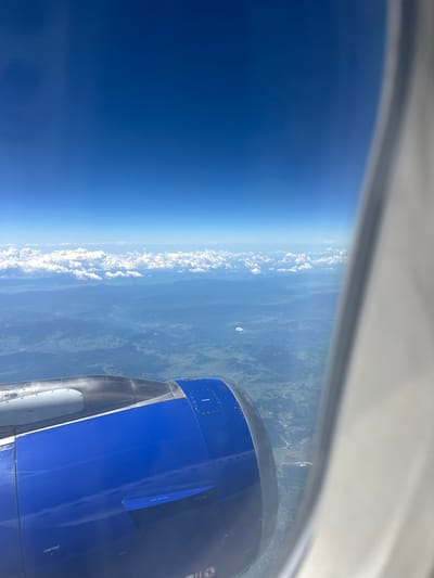 The image is a photograph taken from an airplane window, showing a view of the earth from above. In the foreground, a portion of a blue and silver airplane engine is visible, extending into the lower left corner of the frame. The engine casing is metallic and appears to have some markings and rivets.

Above the engine, the earth's surface is visible, appearing as a patchwork of green and brown terrain with hints of scattered buildings or infrastructure. There are rolling hills and possibly some bodies of water. A line of white clouds stretches across the middle ground, obscuring parts of the landscape below.

The sky is a vibrant, clear blue, dominating the upper portion of the image. A bright white haze or glare is present along the right edge of the frame, which is likely the airplane window frame or glare from the sun.

The question asks for the location in the city of Viehhausen, Austria. However, there are no explicit visual cues within the image that definitively place it in Viehhausen, Austria, or any specific city. The landscape is generic and could be from various regions. Without further information or context, it is not possible to confirm the location.