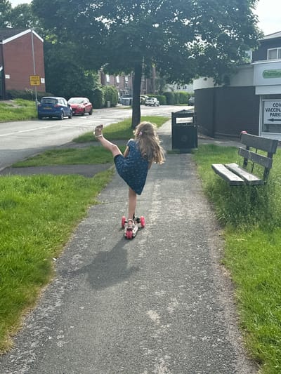 A young girl on roller skates is on a sidewalk, kicking with one leg. The location is on a sidewalk in Macclesfield, United Kingdom. The sidewalk is surrounded by grass, a bench, and a trash can. Behind the sidewalk, there is a pharmacy (Andrew's Pharmacy) and a sign pointing towards a COVID-19 vaccination center. Further back, there is a street with parked cars.