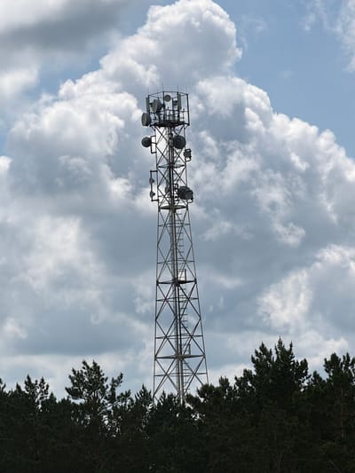A tall, lattice communication tower is centered in the frame, extending from just above a dense line of dark green pine trees to a partly cloudy sky. The tower is made of metal supports and has several antennas and satellite dishes mounted at various levels. A ladder runs vertically along one side of the tower. The trees form a dark, textured band across the bottom of the image. The sky above is a mix of bright blue and large, fluffy white and gray clouds. The scene appears to be located in an outdoor, possibly rural or semi-rural, area. There are no people or animals visible in the image. The setting is consistent with a location in or around the city of Tsiolkovsky, Russia, given the presence of such infrastructure and coniferous forests common to the region.