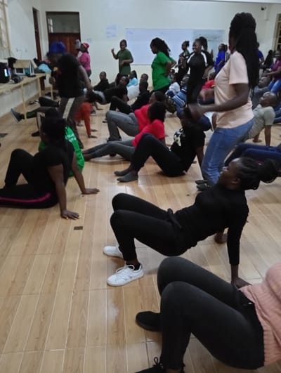 This image captures a lively indoor exercise or dance session involving a diverse group of individuals, predominantly young adults of African descent. The activity takes place in a spacious room with light-colored walls and a distinctive light brown tiled floor, designed to resemble wooden planks.
Many participants are actively engaged in a physical exercise, with several performing a "crab walk" or "reverse plank" position, supporting themselves on their hands and feet with their torsos elevated. In the foreground, a woman dressed in all black demonstrates this pose. Other individuals are seen standing, moving, or preparing for the exercise. They are dressed in casual athletic wear, including t-shirts, jeans, and track pants, with some wearing sneakers and others barefoot or in socks. Hair styles such as braids and dreadlocks are visible among the participants.
The general mood appears energetic and focused, with individuals showing concentration and engagement. On the far left, a person wears a bright green shirt with the partially visible text "I DO." In the background, a whiteboard is mounted on the wall, although any writing on it is illegible. A wooden bench or table with some electronic devices, possibly for music, is present along the left wall. The lighting in the room is bright, suggesting either daylight from unseen windows or artificial illumination. The setting is consistent with a community hall, gym, or multipurpose space located in Riara Ridge, Kenya.