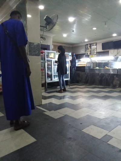 The image shows an interior scene with a person wearing a blue robe standing in the foreground. The person is looking at the camera, and their right foot is partially visible. The person is standing on a tiled floor with a black and white zig-zag pattern. The background of the image shows a woman standing in front of a glass-fronted display case. The woman is wearing a black jacket, blue jeans and appears to be looking at something in the display case. To the right of the display case is a counter with a display case containing some food. There is also a man in the image, wearing a light blue jacket, who is standing near the counter and looking at the woman in the black jacket. The man's right hand is visible and is holding a small brown bag. There are also a number of ceiling lights, a ceiling fan, and a green tiled wall in the image. The scene is located in a restaurant in Ovwian, Nigeria. The coordinates are 5.503 latitude and 5.799 longitude.