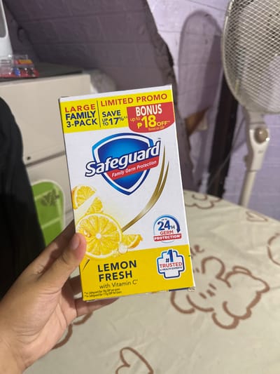 A person's hand is holding a box of Safeguard Lemon Fresh bar soap with Vitamin C. The box is yellow and white with images of lemons. Text on the box reads "LARGE FAMILY 3-PACK", "LIMITED PROMO", "SAVE UP TO 17%", "BONUS P18 OFF", and "24H GERM Protection". There is also a seal that says "#1 TRUSTED BY HEALTH EXPERTS". The background is blurry and appears to be an indoor setting with a fan and a table with a patterned cloth. The lighting suggests it is daytime. The location is not explicitly stated as Manila, Philippines.