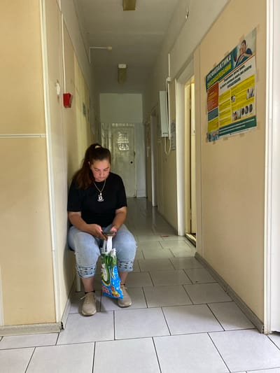 A woman sits on a stool in a hospital hallway, looking down at her phone. She is wearing a black t-shirt, light blue jeans, and beige sneakers, and she has a white bag on her lap. The hallway is lined with cream-colored walls and pale gray tiled floors. A poster about cancer prevention is mounted on the wall to the right of the woman. A red fire alarm box is visible on the wall to the left. The lighting appears to be fluorescent, originating from ceiling fixtures. The scene suggests a waiting area in a medical facility.