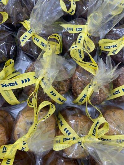 The scene, located indoors in Nes Ziona, Israel, presents a close-up, overhead view of multiple individually packaged baked goods. Each item, resembling a muffin or small cake, is sealed within a clear, transparent plastic bag. Each bag is secured with a bright yellow fabric ribbon tied into a bow. The ribbon features repeating dark blue text, specifically the Hebrew word "איקאה" (IKEA), and elements of the IKEA logo. The baked goods exhibit variations in color and texture, including darker, dense brown items and lighter, possibly oat-based items with visible inclusions. They are arranged compactly on a light-colored surface, suggesting placement within a container or on a display. No persons or overt actions are depicted.