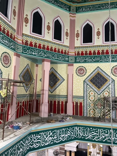 This image captures the richly decorated interior of a building, likely a mosque, in Bale, Indonesia, appearing to be in the final stages of renovation or construction. The walls and ceiling are adorned with elaborate Islamic art and calligraphy.
Dominating the scene are wide bands of green featuring intricate white Arabic calligraphy, which appear to run along the upper sections of the walls. Below these, sections of geometric patterns in red and brown hues add further detail. The main wall and ceiling surfaces are painted a light cream or beige, featuring subtle, repeating geometric patterns in a lighter tone.
Multiple arched windows with dark panes are visible along the upper walls. In the ceiling areas, diamond-shaped windows or vents with textured glass are set within intricate square borders of green, gold, and white. Circular medallions with Arabic script are also incorporated into the ceiling and wall designs.
The overall color palette is vibrant, comprising greens, reds, creams, gold, and light pink columns or supports. Metal scaffolding is present in the lower right and central parts of the image, alongside what appears to be construction debris, suggesting ongoing work. The intricate details and vibrant colors showcase a blend of traditional Islamic aesthetics and modern craftsmanship. The lighting suggests an indoor setting, possibly illuminated by artificial lights.