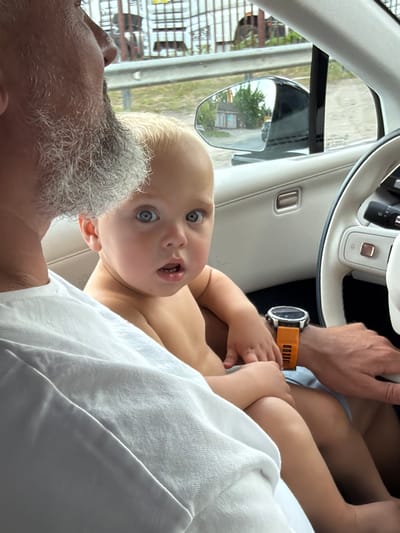 An adult, with a gray beard and wearing a white shirt, is seated in the driver's side of a car. They are holding a baby, who is naked and has blonde hair and blue eyes, on their lap. The baby is looking directly at the camera with a surprised expression. The interior of the car is visible, including the steering wheel, dashboard, and a side mirror reflecting an outdoor scene with buildings and trees. The exterior environment appears to be a road or parking lot, with hints of industrial or commercial structures in the background, consistent with a location like Aprelevka, Russia. The lighting suggests it is daytime.