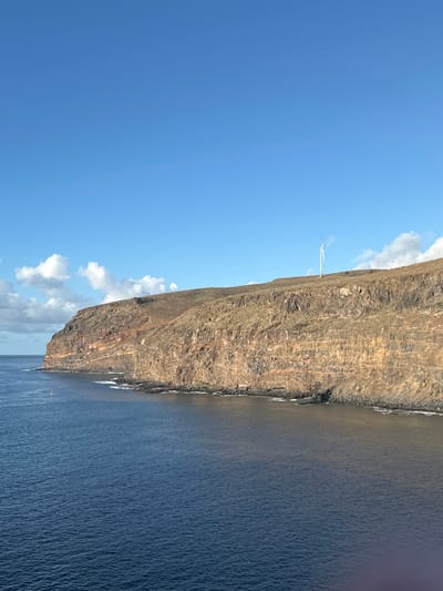 This image depicts a coastal landscape in San Sebastián de la Gomera, La Gomera, Canary Islands, Spain. The scene is dominated by a vast expanse of deep blue ocean in the foreground and midground, meeting a prominent, steep cliff face that extends from the mid-right across the horizon to the left.
The sky occupies the upper half of the frame, a clear, bright blue with scattered white cumulus clouds, primarily concentrated towards the left horizon. The ocean water is calm with minor surface ripples.
The landmass features a massive cliff, characterized by horizontal geological stratification in shades of reddish-brown and dark grey rock. The base of the cliff meets the sea with a rocky shoreline. Above the cliff, the terrain slopes upwards, covered in sparse, dry, brown-green vegetation, with some terraced sections visible. A single, tall, white wind turbine with three blades stands on this upper, vegetated slope of the cliff in the mid-right portion of the image. A small, red-orange conical buoy is visible floating in the ocean in the middle-left, near the base of the distant coastline. No people or specific actions are depicted in the scene.