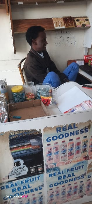 A man sits inside a small shop, visible from the torso up. He wears a brown jacket and blue jeans. Behind him, the wall has shelves displaying rectangular desserts. To his right, behind a white countertop filled with various jars, are aging posters promoting "REAL FRUIT REAL GOODNESS," and an ad with "ENTER" and "23804" visible. The countertop surface is also littered with a white notepad. Handwritten numbers are visible on the wall behind the man.