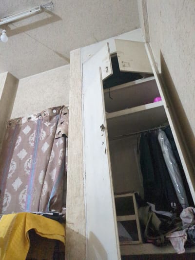The image shows an indoor scene, likely a room. A large, open cabinet structure dominates the upper part of the frame, with various items stored inside, including plastic-wrapped objects and a bag. The cabinet appears to be made of light-colored wood or laminate. Below the cabinet, a patterned fabric, possibly a curtain or draped material, hangs down. The fabric has a repeating floral or paisley-like motif in shades of brown and cream, with some iridescent blue and pink streaks. A yellow cloth is partially visible to the right of the fabric. The walls in the background are textured and a pale beige color. In the lower left corner, a fluorescent light fixture is mounted vertically on the wall, with a white lightbulb attached. The overall impression is of a storage area or a room with functional, utilitarian elements. The lighting suggests it is daytime, but no specific time or weather cues are apparent. No text is visible in the image.