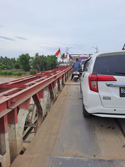 A white SUV is stopped on a bridge in Tingkeummanyang, Indonesia, facing forward. The bridge has red metal railings and a dirt and metal surface. A man on a white scooter is in front of the SUV. Indonesian flags are visible on poles along the bridge. Lush green trees and vegetation are on the left bank of the river beside the bridge, with some houses visible among the trees. The sky is overcast.