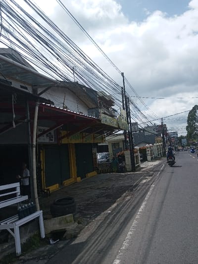 This image depicts a bustling commercial street scene. While identified by the user as Gubbisamamidi, India, the visible signage, specifically "BAKSO SOPU" and "BENGKEL" (workshop), along with the architecture, strongly indicates an Indonesian location.
The scene is under an overcast sky with white and grey clouds. A dense network of numerous black electrical and communication cables crisscrosses the upper portion of the frame, strung between multiple utility poles lining the street.
On the left side of the street, a row of commercial buildings is visible. The closest structure features a red corrugated metal awning supported by poles. Partially visible beneath this awning is a sign that reads "BENGKEL." Adjacent to it is a storefront with yellow trim and two closed dark green roll-down shutters, displaying a prominent sign for "BAKSO SOPU." In the immediate foreground on the left, a white metal shelving unit holds several dark-colored bottles, possibly lubricants. Inside the open-fronted shop to the far left, an older man in a light-colored shirt is visible, appearing to handle an object. On the ground near the storefront, two black tires are stacked horizontally next to a concrete block. Further down the sidewalk, two individuals, one wearing a head covering, are seated or crouching near a low, decorative white wall.
The asphalt road, marked by a worn white lane line, shows a person riding a dark motorcycle, wearing a helmet and dark attire, traveling away from the camera. Other vehicles, including motorcycles and cars, are visible further down the road in the distance. The right side of the road is bordered by a series of white, decorative pillars or fence sections, with tall trees visible further down the street on the right.
