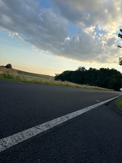 An eye-level, low-angle shot captures a paved road with a white dashed line running along its surface, curving to the right in the foreground. The road is dark gray asphalt, textured with small pebbles. Beyond the road, to the left, a gently sloping field of golden-brown crops stretches into the distance, bordered by tall, dry grass. Further back, a dense line of dark green trees forms a forest edge. Above, the sky is a mix of pale blue and dramatic, heavy gray clouds, with patches of sunlight breaking through, creating a soft, diffused light. The scene appears to be a rural setting, possibly on the outskirts of Beltershain, Germany, given the presence of agricultural land and a forest. There are no people or animals visible in the image.