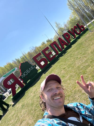 A medium shot captures a man in Крошынскі сельскі Савет, Belarus, posing in front of a sign spelling "БЕЛАГРУСЬ" (Belarus). The man is angled towards the camera, making a peace sign with his right hand. He wears a blue and white plaid shirt over a brown t-shirt, with a maroon and white baseball cap atop his head. He has light skin, a mustache, and a wide smile displaying his teeth. The sign, featuring large red letters with a green border, is prominent in the foreground. A small green sign with white Cyrillic text is visible below. The background shows a sunny day with blue sky, trees with budding green leaves, and a grassy field. Tall metal poles stretch into the sky on the left side of the frame.