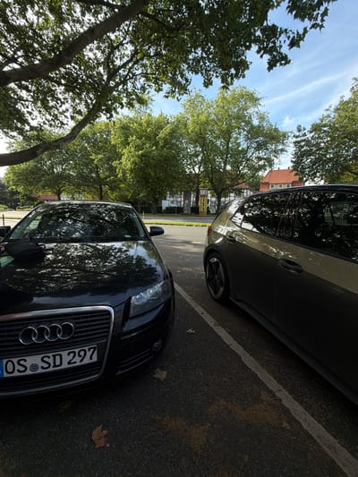Two parked cars are visible from a low angle in a tree-lined urban area. The car on the left is a black Audi with a license plate that reads "OS SD 297". The car on the right is a two-toned vehicle, possibly an electric car, with a grey and gold exterior. The background shows a street with buildings and trees. Based on the license plate format and the architecture visible, this scene is likely in or around Herford, Germany. The image is taken during the daytime, with sunlight filtering through the leaves of the trees, creating dappled light on the cars and the asphalt. There are no people visible in the image.
