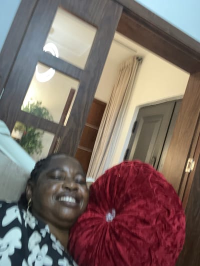 A medium shot, taken from an elevated angle, features a woman smiling widely with a red, circular cushion resting beside her head. The woman is wearing a black and white floral top, and gold jewelry is visible near her ear. The background includes a partially visible wooden framed doorway or window, with reflective panels. A plant is visible through one of the panels. Above the doorway, there is a ceiling light or window with light-colored blinds partially drawn. The image does not reveal any information regarding its location in the city of Mopo-Akinlade, Nigeria.