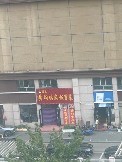The image shows a street scene in Manzhouli, China, featuring storefronts and parked vehicles. The buildings are modern, with large windows and signage. One prominent sign in red and gold lettering reads "黄焖鸡米饭" (Huang Men Ji Mi Fan), indicating a restaurant specializing in braised chicken and rice. Next to it, a blue and white sign displays "CAI N IAO 菜鸟驿站" (Cai Niao Post Station), a logistics service. Several cars are parked along the street, including a silver SUV and a black sedan. In front of the buildings, there are trees and some sidewalk activity, with a person visible inside the Cai Niao station. The overall impression is a typical urban commercial area.