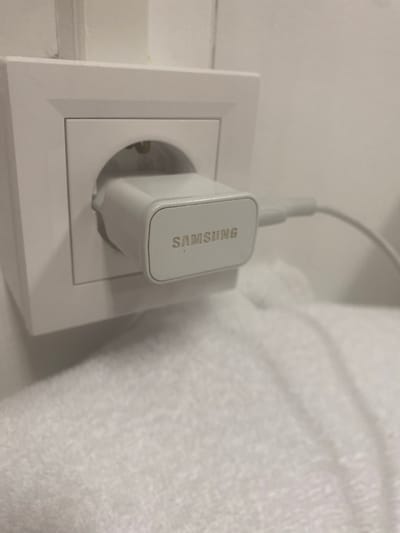 A white Samsung brand phone charger is plugged into a white electrical outlet on a light-colored wall. A white charging cable extends from the charger and disappears out of frame to the right. The outlet is set into a white wall plate, which is slightly recessed into the wall. Below the outlet, a textured white surface, possibly a towel or blanket, is visible. The scene is indoors, and the lighting is soft and diffused. The image is cropped to focus on the outlet and charger.