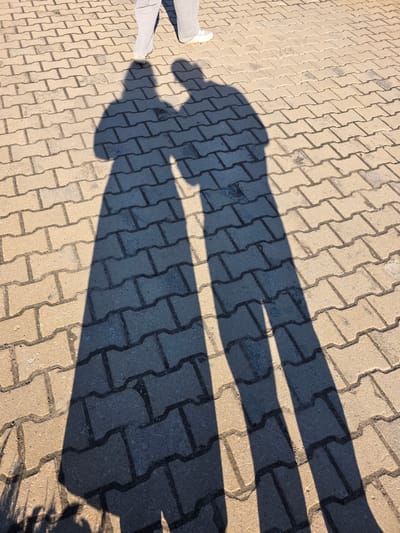 The image shows the shadow of a person standing on a brick-paved surface. The person is wearing light-colored pants and shoes. The shadow is elongated, suggesting a low sun angle. The brick pavers are light brown. The location, Repino, Russia, is not discernible from the image itself. No other people, objects, or interactions are present. The environment appears to be outdoors.