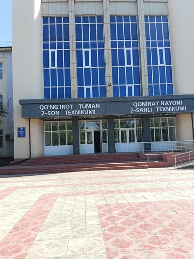 This is a frontal exterior shot of a modern building in Kungrad, Uzbekistan, featuring large blue-tinted windows with white framing on the upper floors and glass doors with white framing on the ground level. Above the entrance, a dark gray sign displays text in white lettering. The building has a light beige facade and is accessed via a wide set of red brick stairs leading to the entrance. A paved walkway with a patterned design of red and beige tiles stretches out in front of the stairs. To the left of the entrance, a blue plaque with crests and text is affixed to the wall. A metal railing system guides people up a ramp to the right of the stairs. The sky is visible in the upper left corner of the image, and some greenery can be seen through the glass doors. The overall impression is one of an institutional or educational facility.